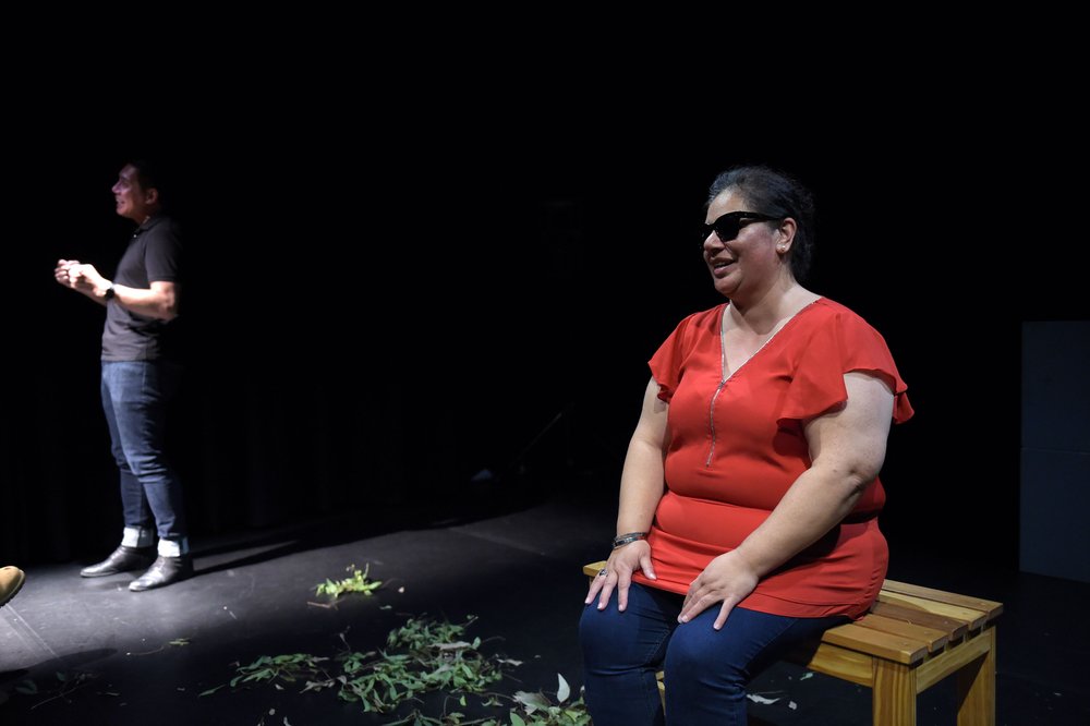  ID: Performer Vanessa Nanai on stage seated at the wooden bench she made, smiling, wearing fire engine red top, blue jeans, and sunglasses, both palms resting on her knees. To her left, standing, is Auslan interpreter Marc Ethan in bright light sign