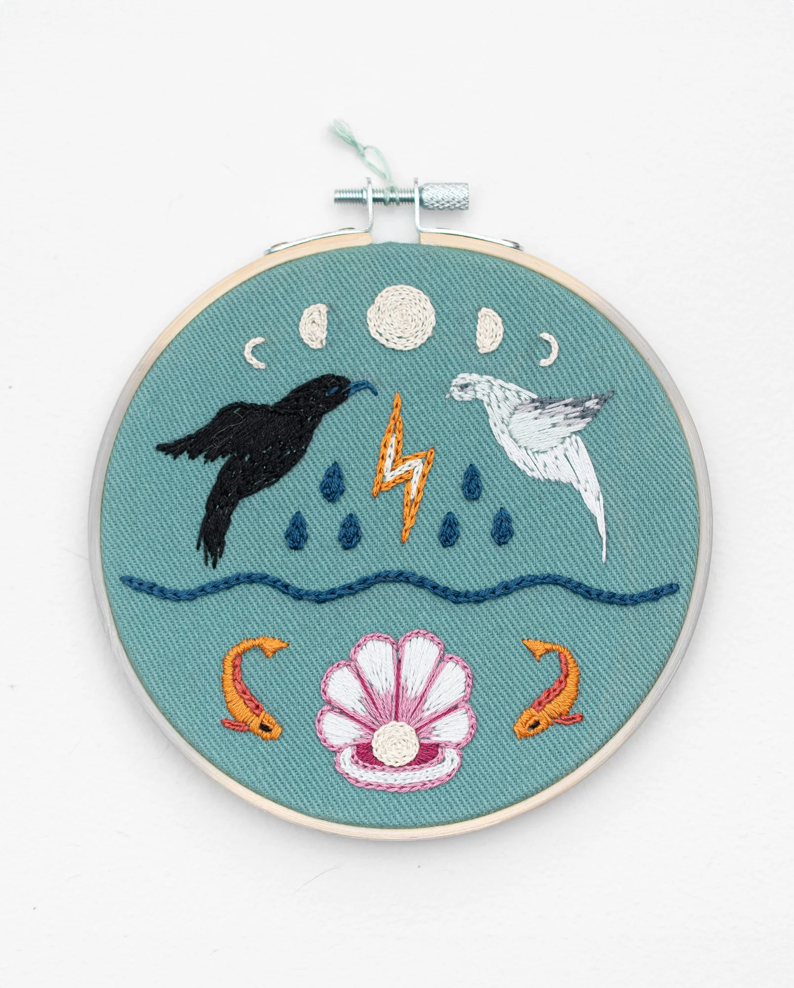 Hand embroidery in a hoop with turquoise fabric depicting totem animals, a crow, a mourning dove, moon cycles, an oyster with a pearl, lightning bolt, and fish