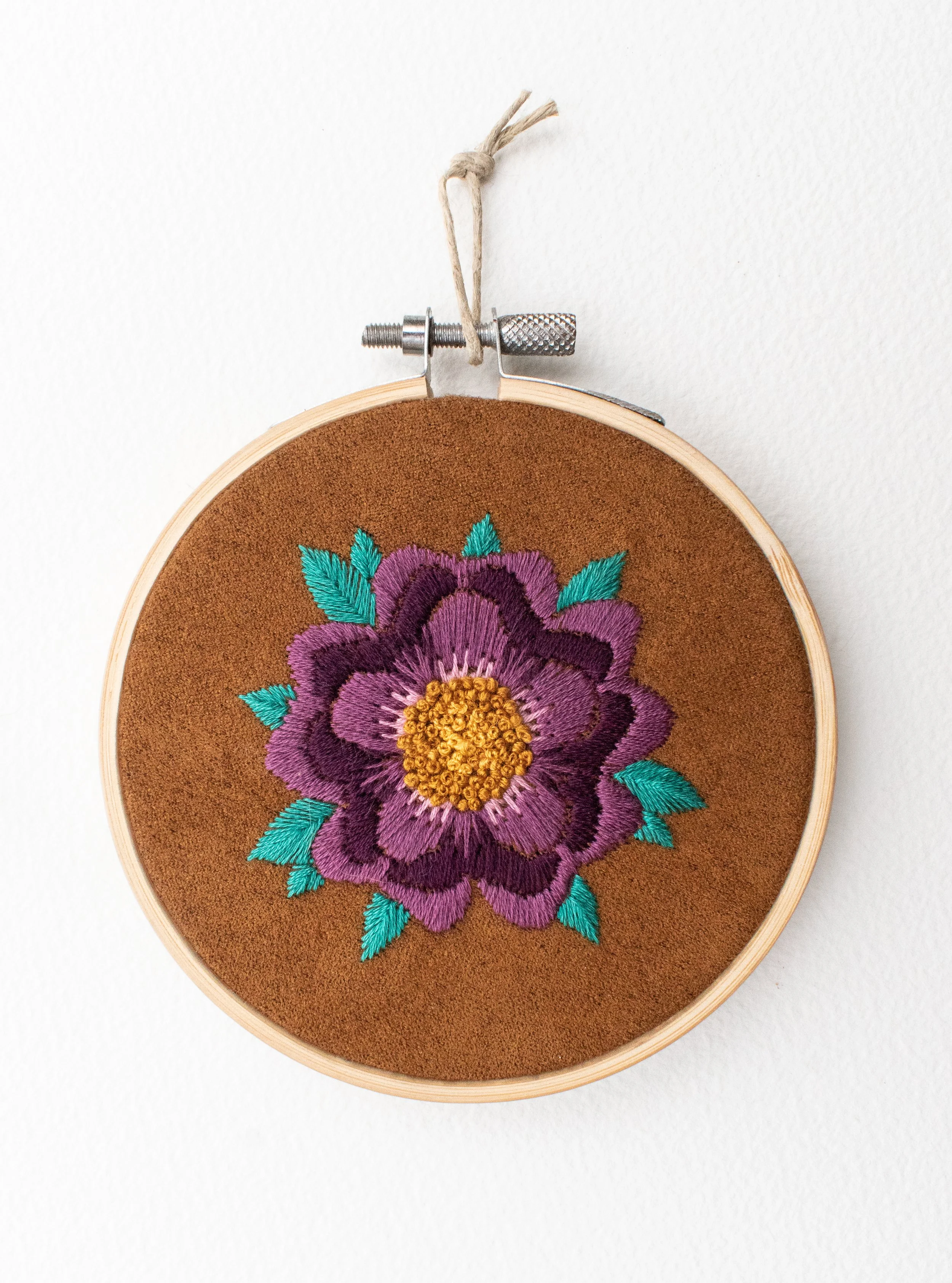 Hand embroidery in a hoop on brown suede fabric depicting a purple hibiscus flower with turqoise leaves