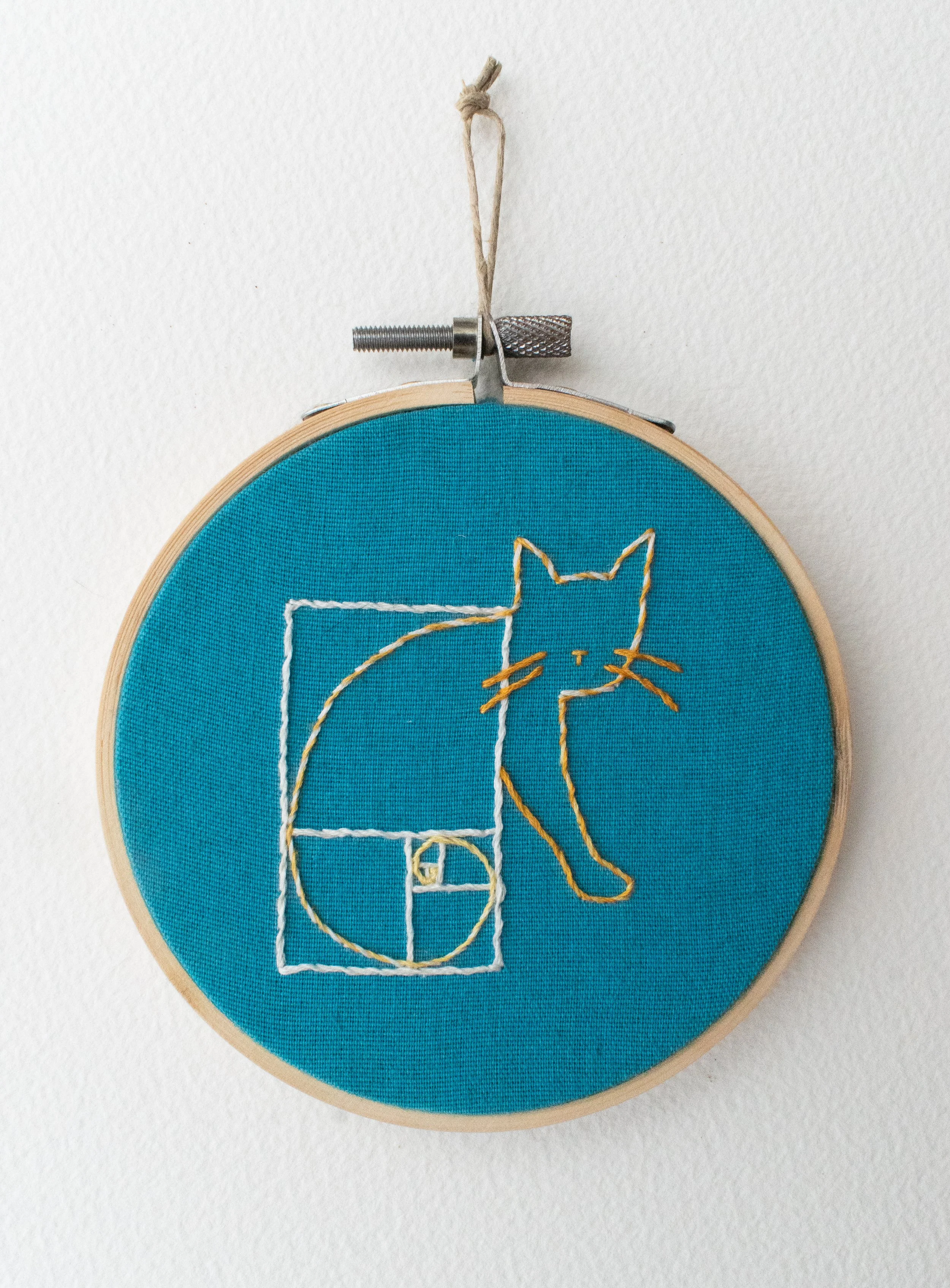 Hand embroidery in a hoop with blue fabric, depicting the outline of a cat whose body and tail align with the fibonacci spiral