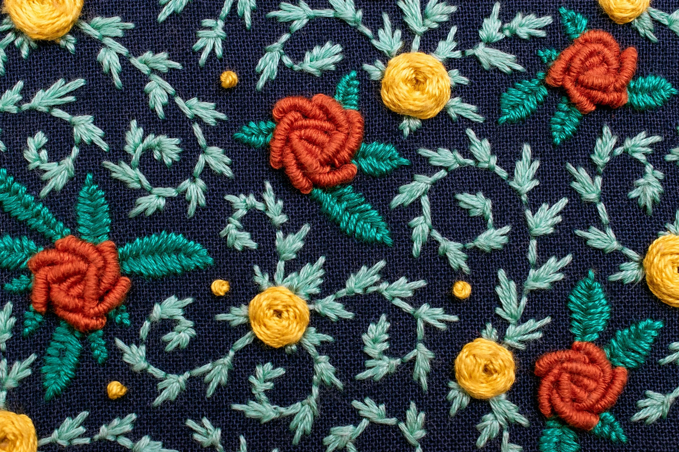 Close-up of hand embroidery with navy fabric, aqua leaves, and orange and yellow flowers forming a pattern