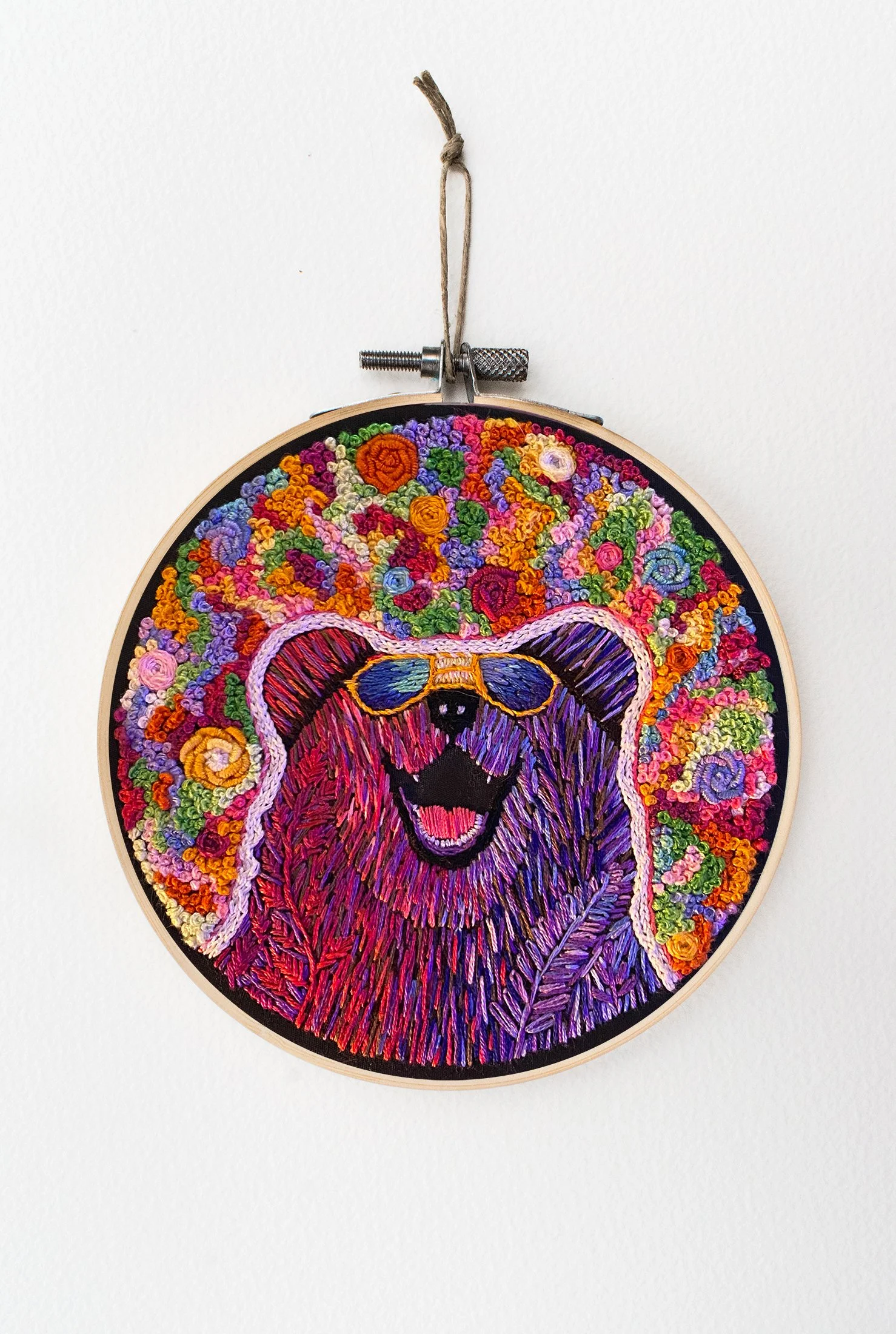Colorful hand embroidery in a hoop depicting a purple and pink bear wearing sunglasses with a background of multi-color flowers covering the fabric