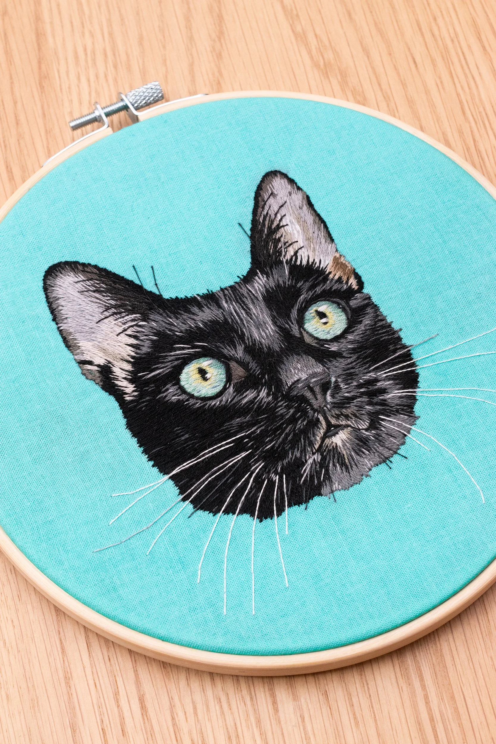 Hand embroidered custom pet portrait of a black cat on turquoise fabric, framed in a hoop, sitting on a wooden table