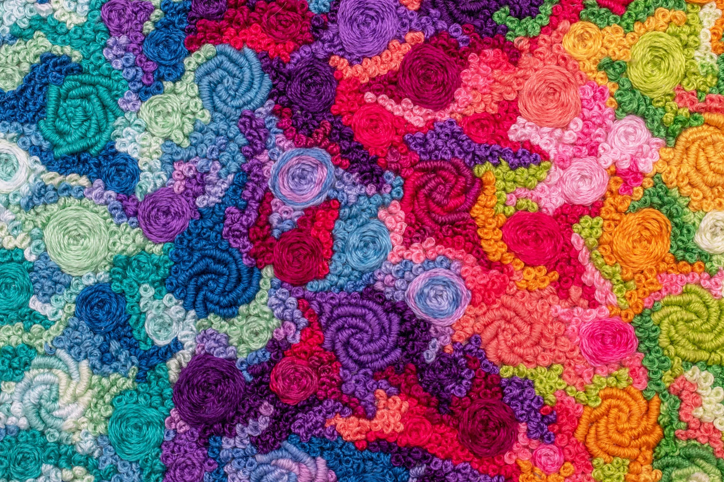 Close-up of colorful rainbow hand embroidery, with bullion roses, pinwheel flowers, and french knots covering the fabric
