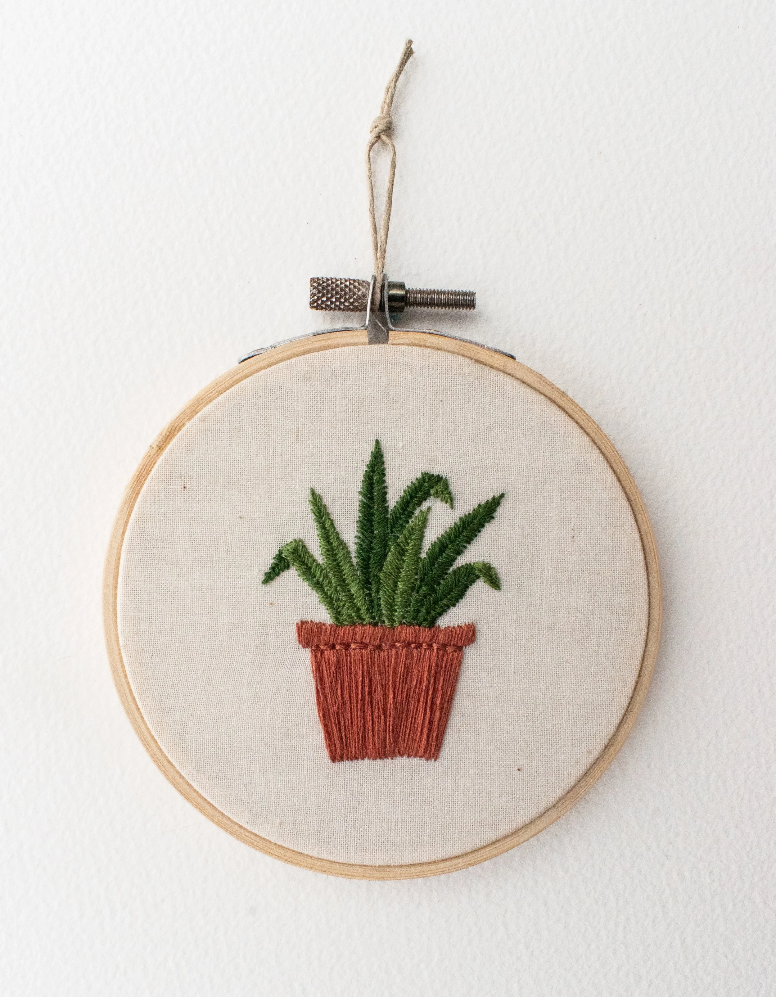 Hand embroidery in a hoop with natural fabric depicting a green leafed plant in a terra cotta pot
