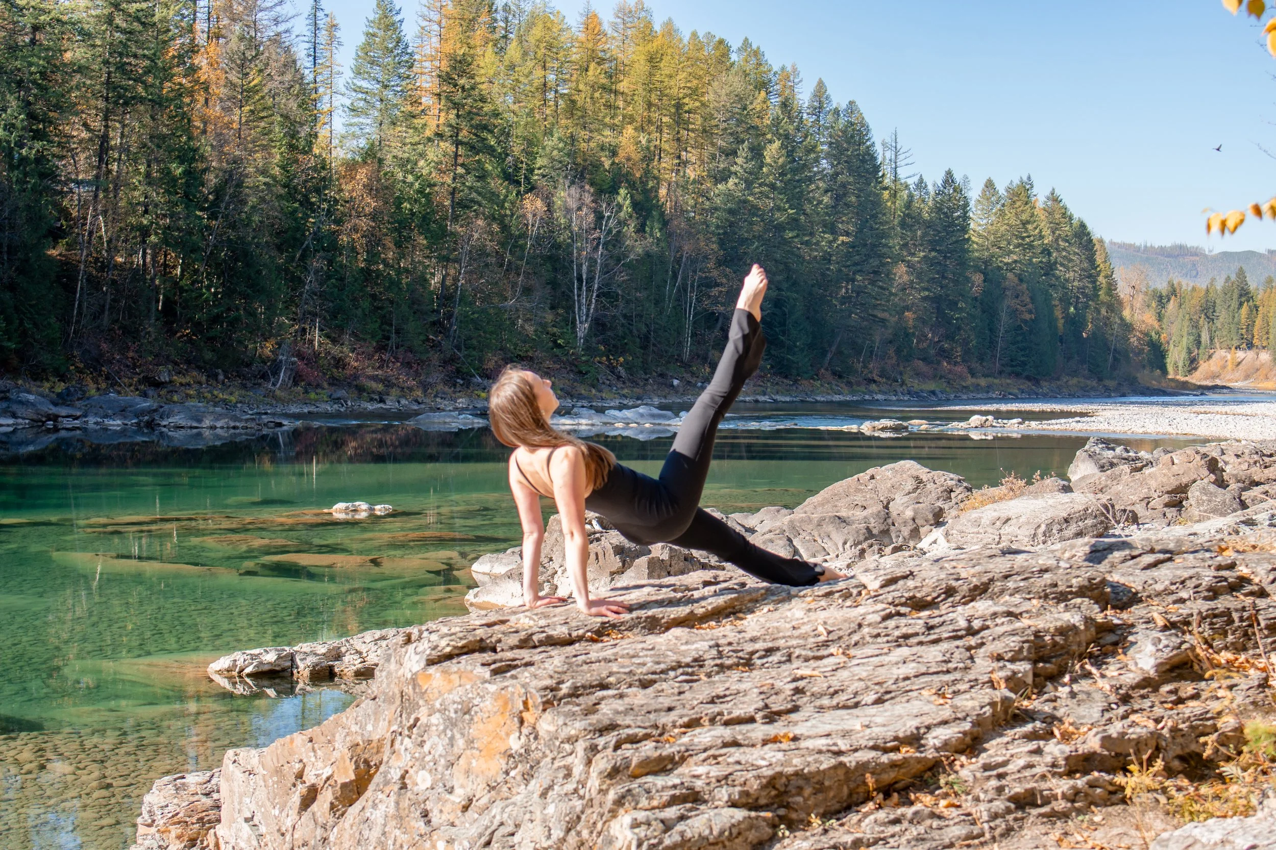 A woman does the Pilates exercise leg pull back on rocks along a river