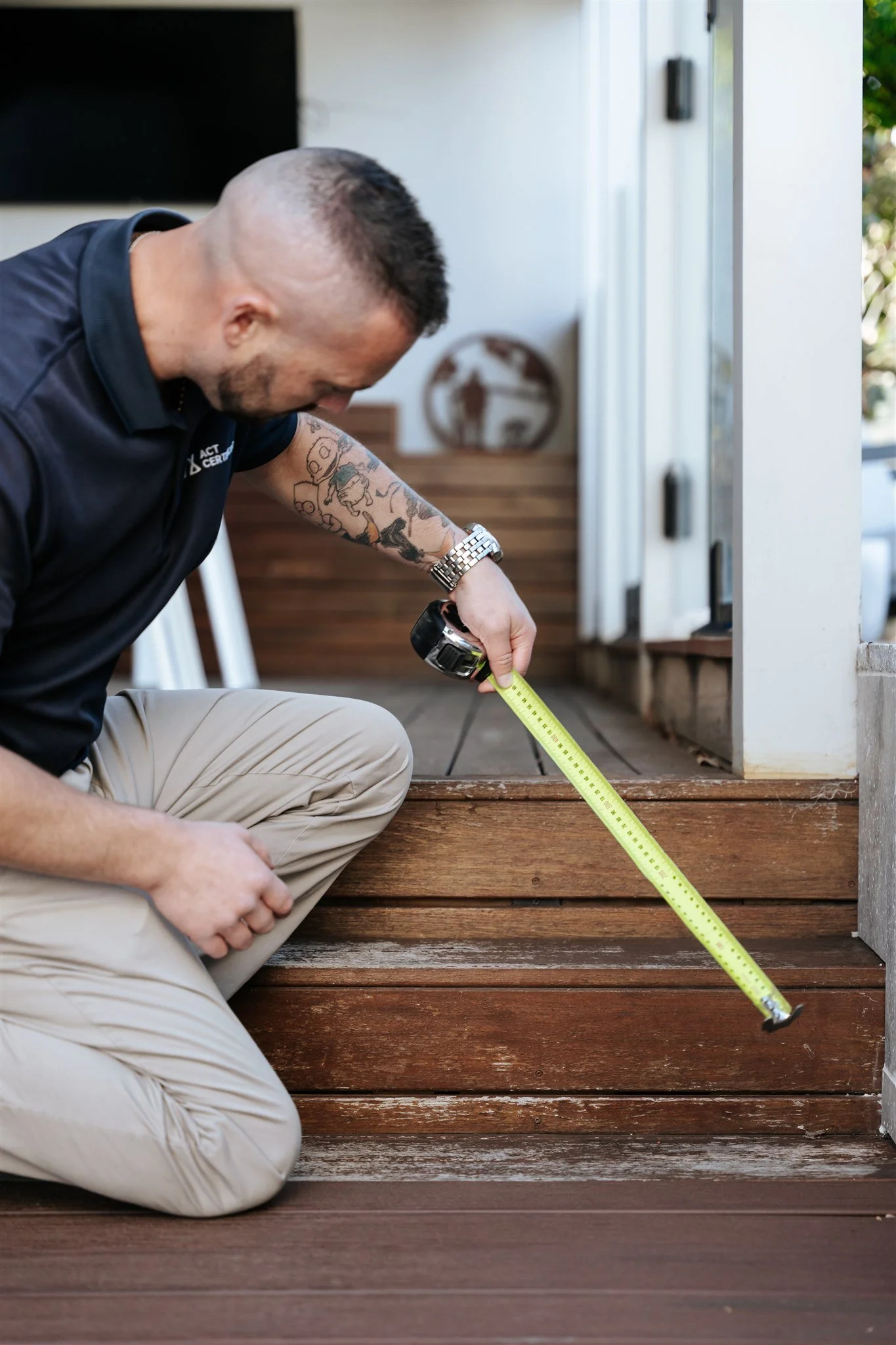 A man kneeling on the wooden stairs outside, measuring the distance with a tape measure.