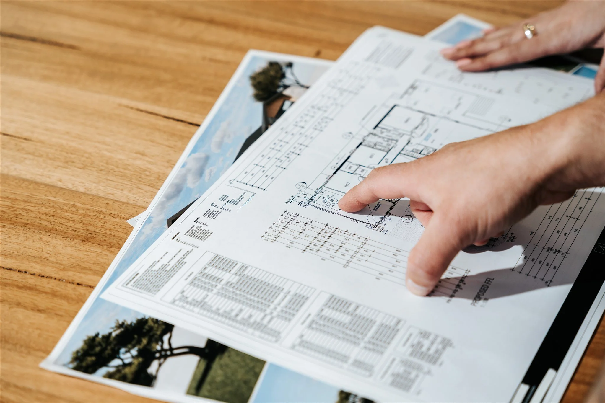 A person's hand pointing at an architectural blueprint on top of a landscape photograph on a wooden table.