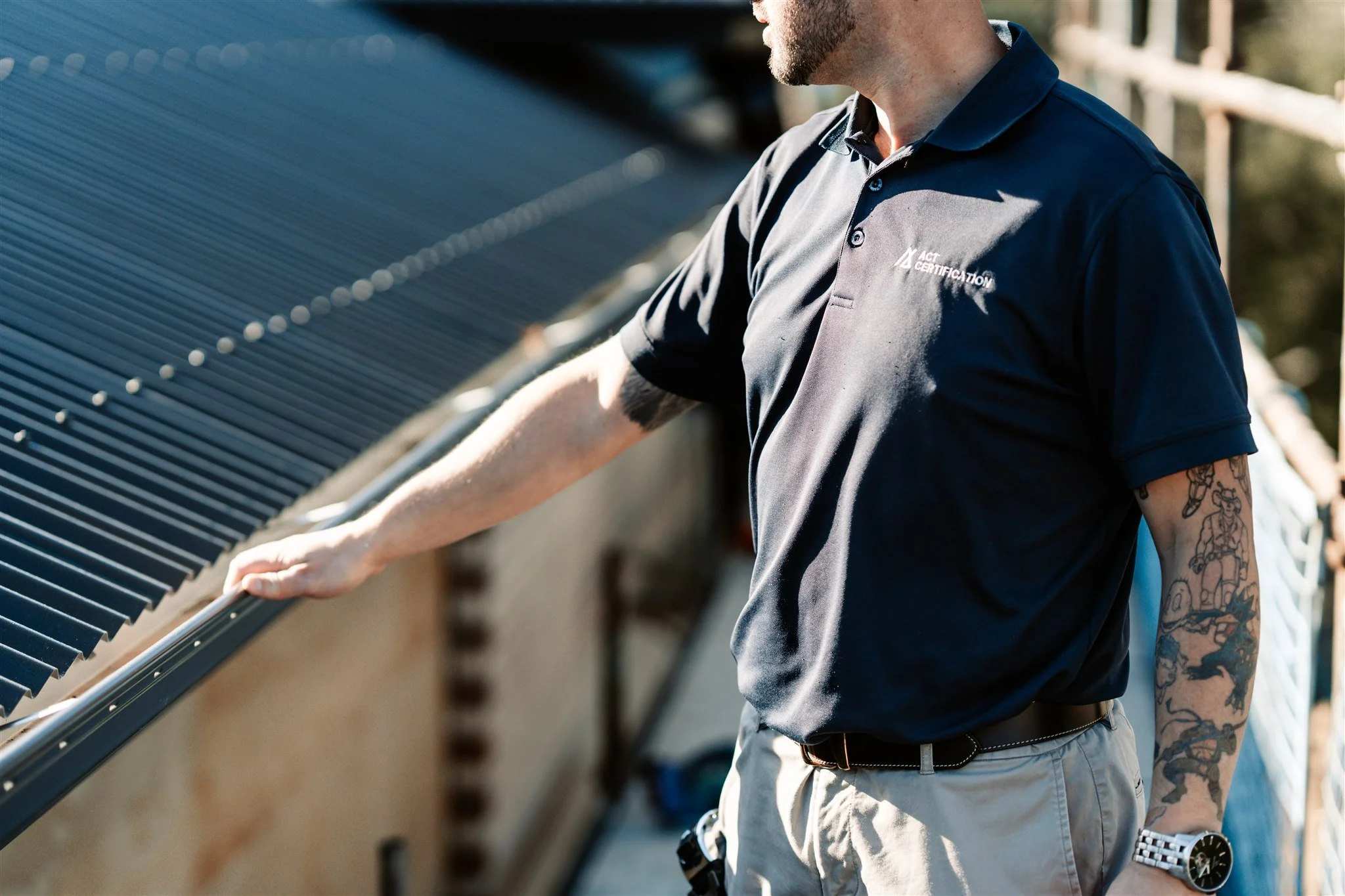 A man inspecting solar panels outdoors, wearing a black polo shirt with certification logo, beige shorts, wristwatch, and tattoos on his arm.