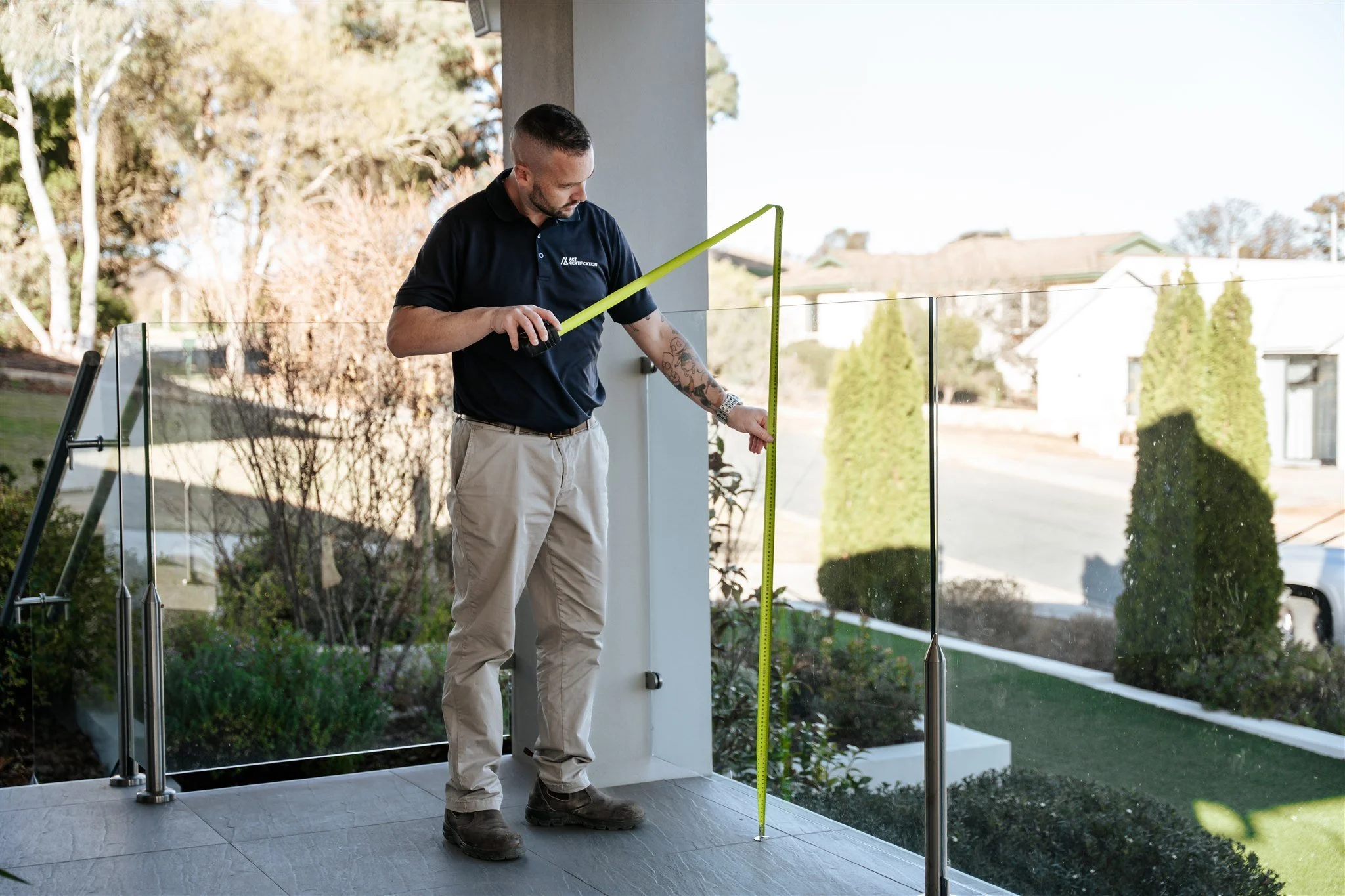 A man is measuring the height of a glass railing on a patio with a tape measure on a clear day.