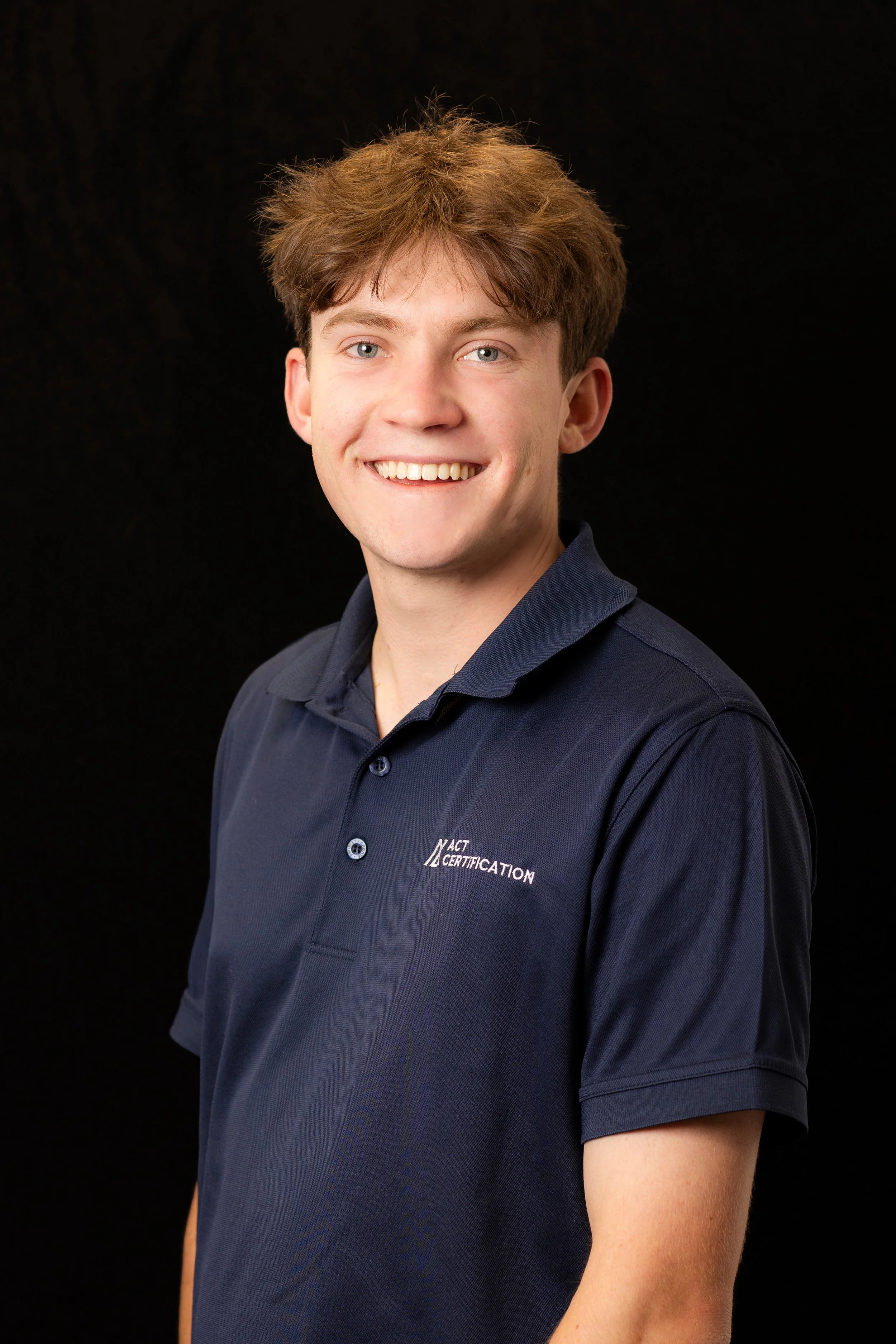 A young man with brown hair, smiling, wearing a navy blue polo shirt with white text 'ACT CERTIFICATION' on the left chest, standing against a black background.