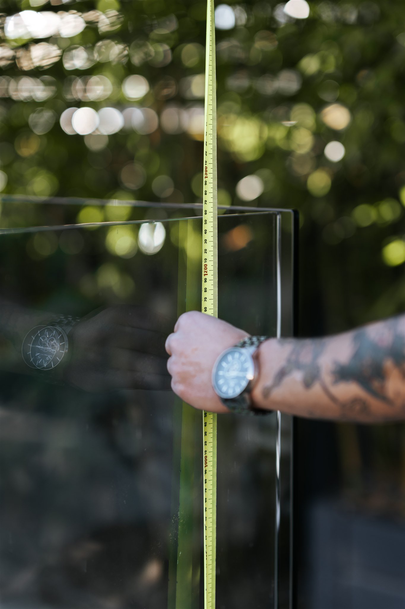 A person's hand, wearing a wristwatch, holds a yellow measuring tape vertically against a glass surface outdoors with a blurred green leafy background.