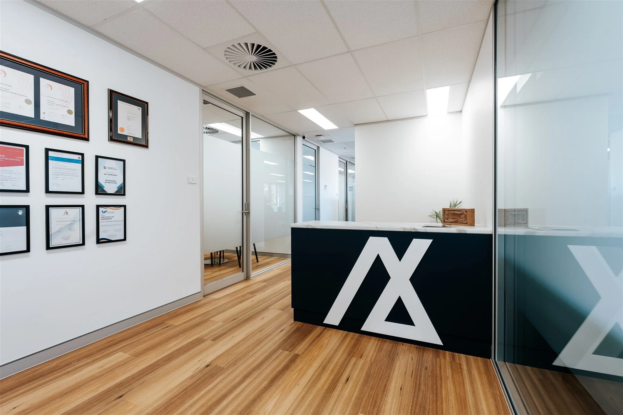 Modern office reception area with a black and white front desk featuring a large Delta symbol, framed certificates on the white wall, glass doors to private rooms, wood flooring, and minimal decor.