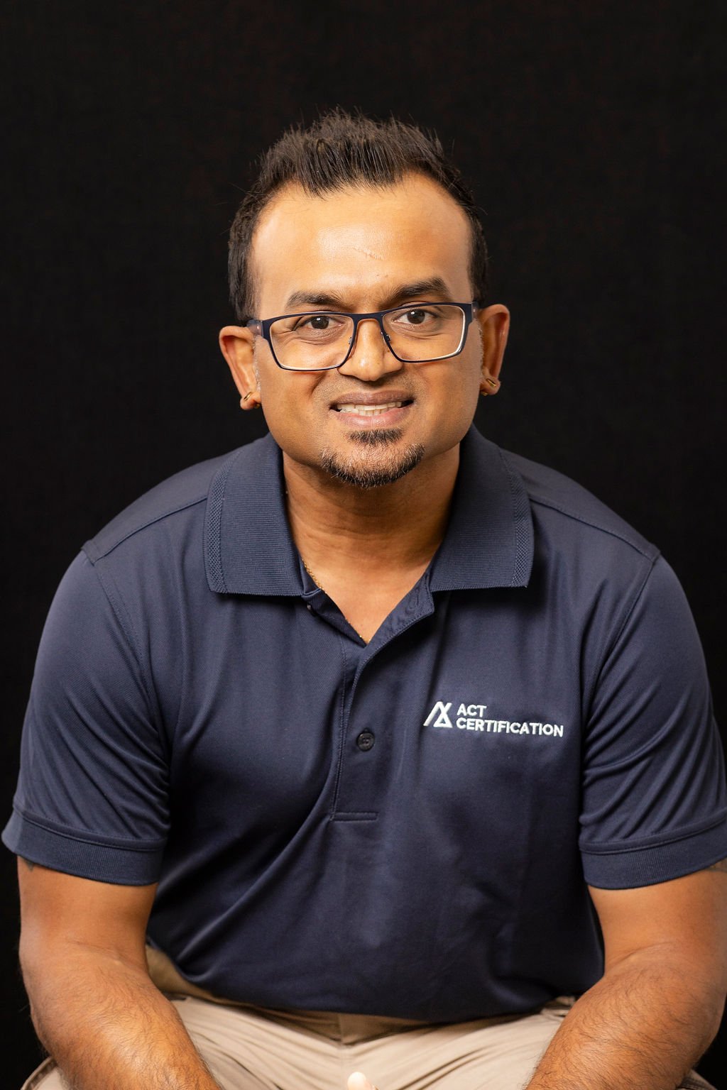 A man wearing glasses and a navy blue polo shirt with ACT Certification logo, sitting against a black background