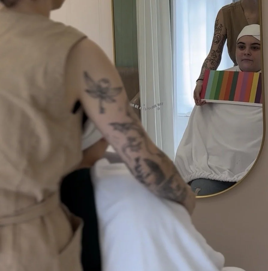 A woman is looking at herself in a mirror while holding a rainbow-colored box, and her reflection shows her with a white head covering and a white dress.
