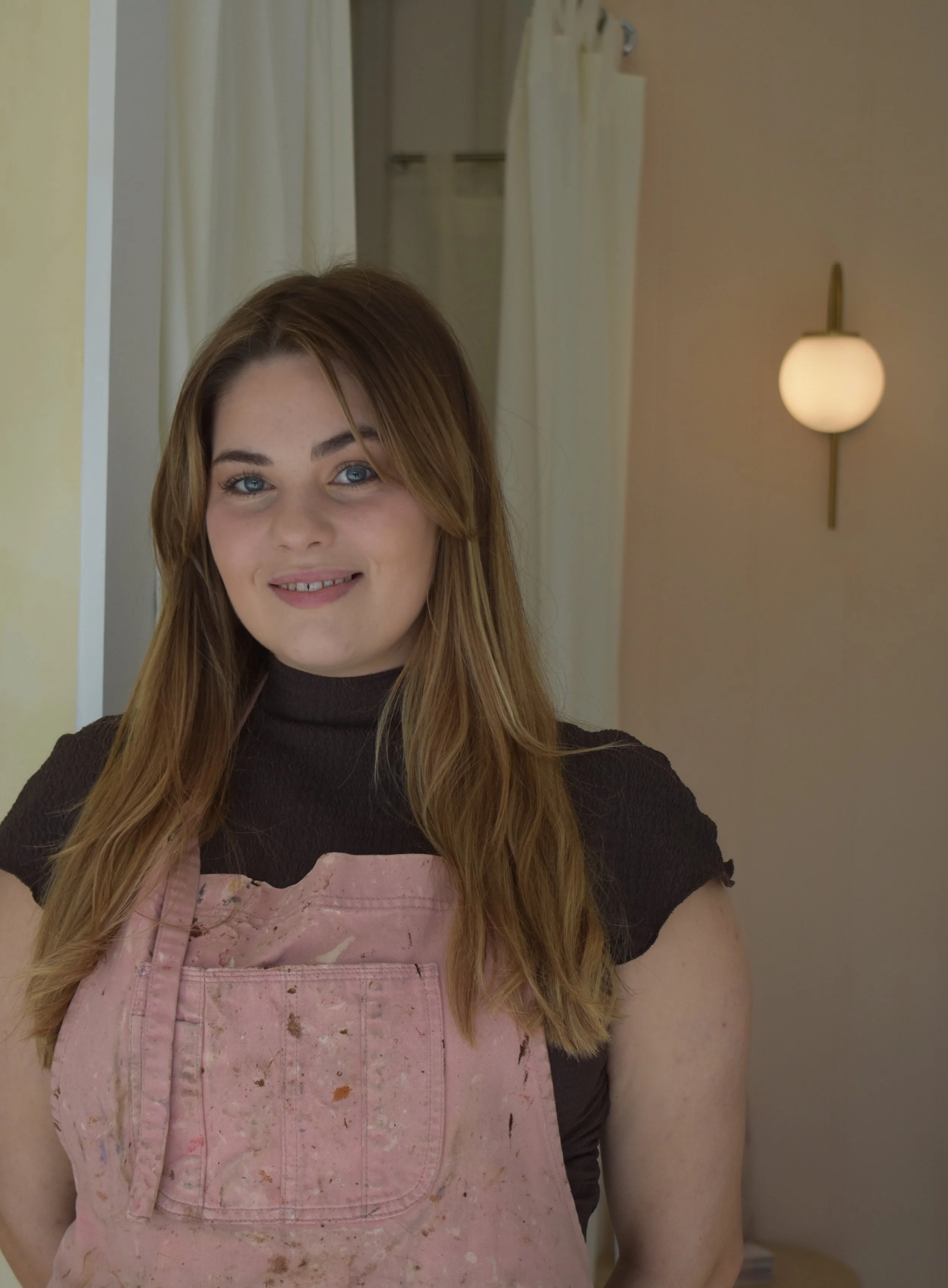 A young woman with long brown hair and blue eyes, wearing a black top and a pink apron, stands indoors with a warm smile. There is a wall sconce with a round white light on the wall behind her, and a doorway with curtains in the background.