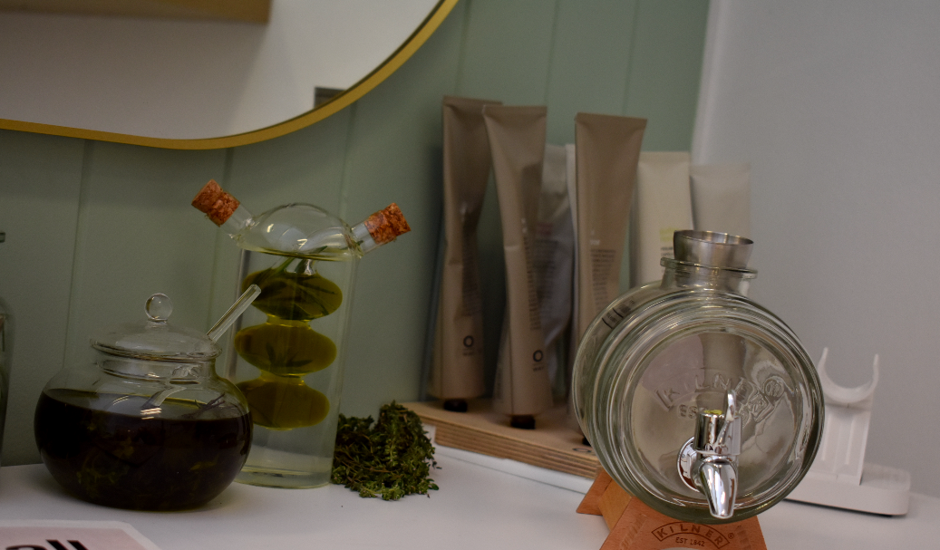 Various bottles and jars containing liquids and herbs on a white surface in front of a mirror and a wall with a green panel.