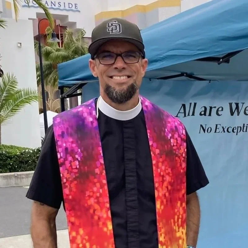 Smiling man wearing glasses, a priest's collar, and a padres hat