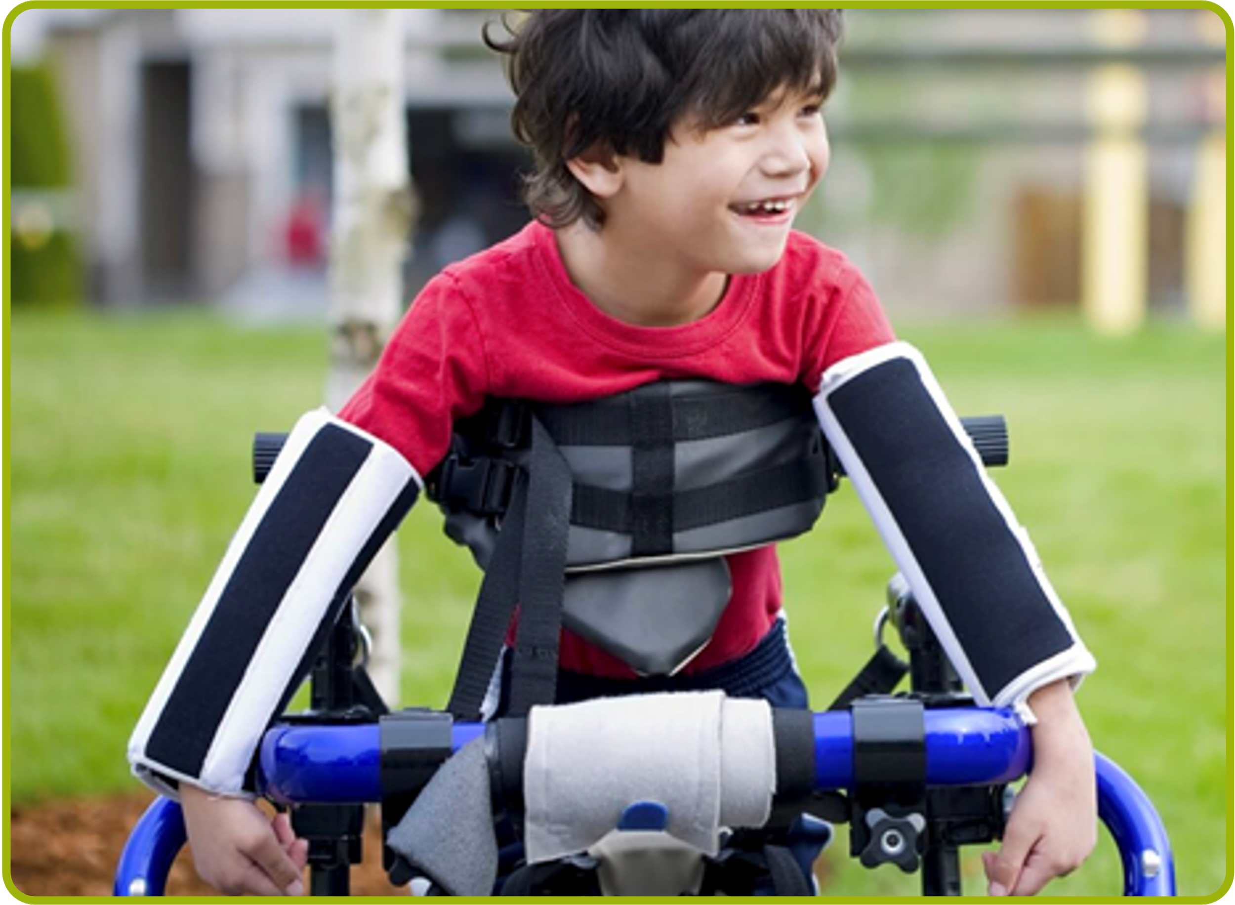 A young boy with dark hair in a red shirt using a wheelchair with elbow immobilizers- harm reduction devices and chest support outdoors on a grassy area, smiling and looking to his left.