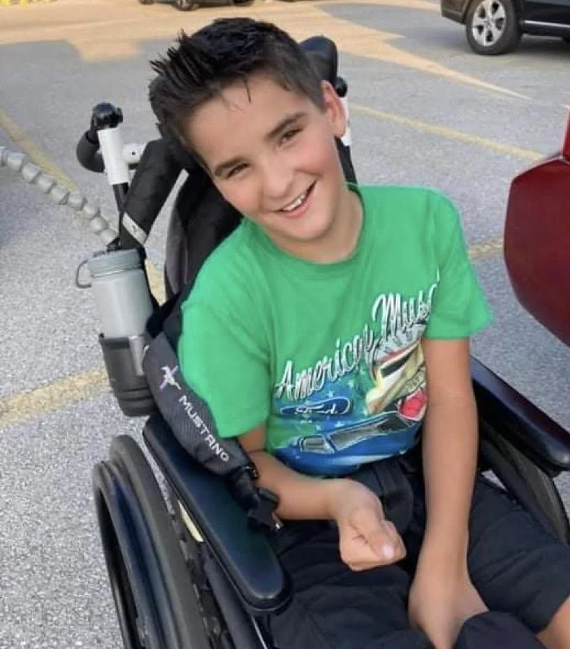 A smiling young boy with dark hair sitting in a power wheelchair on a parking lot, wearing a green T-shirt.