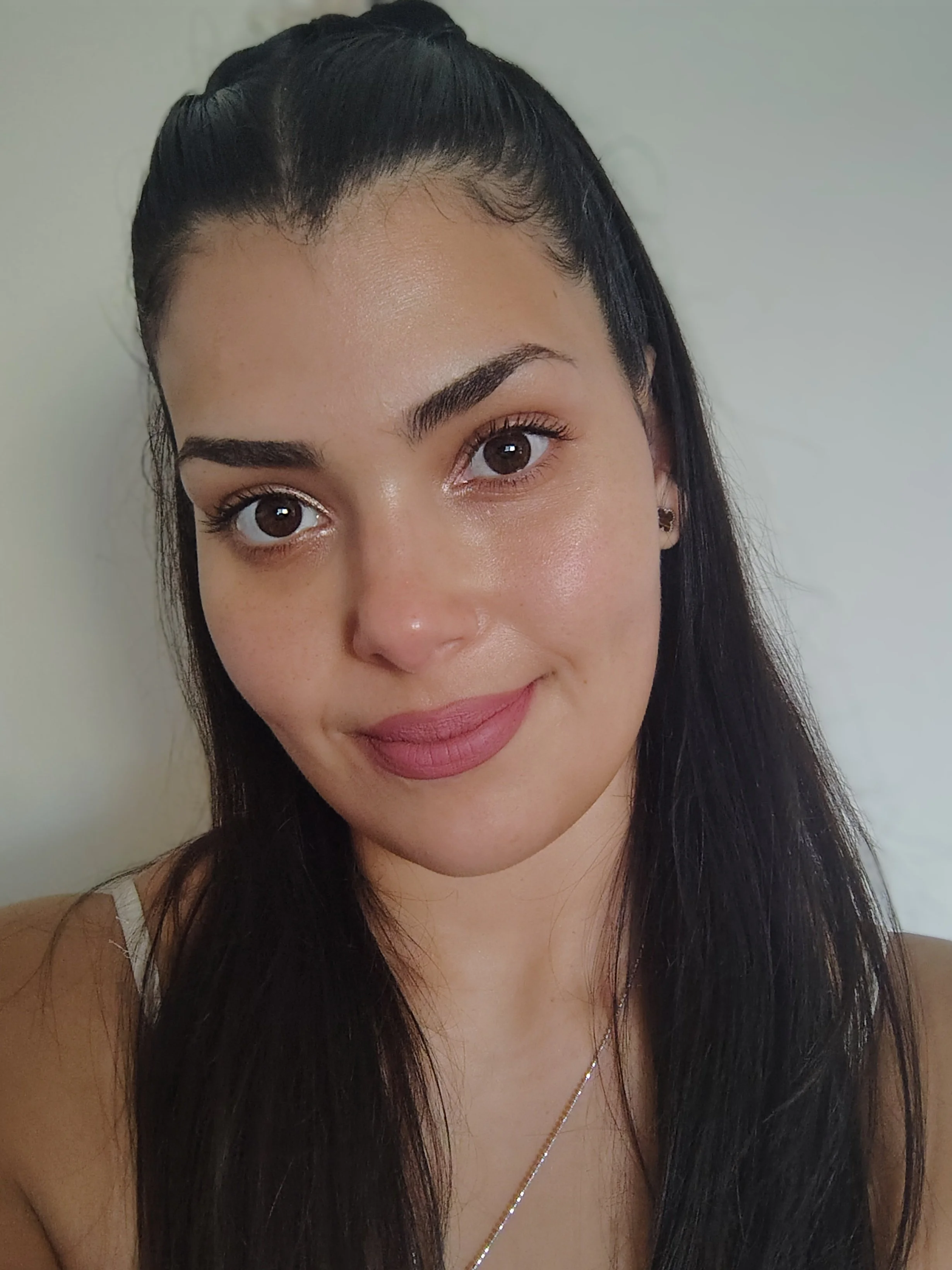 A young woman with long black hair, wearing a light-colored top, a necklace, and small earrings, smiling softly at the camera against a plain light-colored wall.