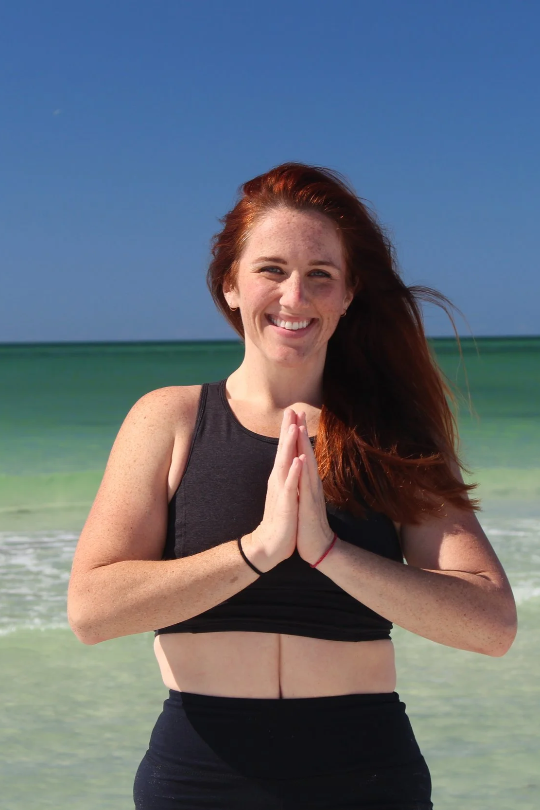 A smiling woman with red hair standing on a beach with ocean waves and clear blue sky in the background, practicing yoga or meditation with hands pressed together.