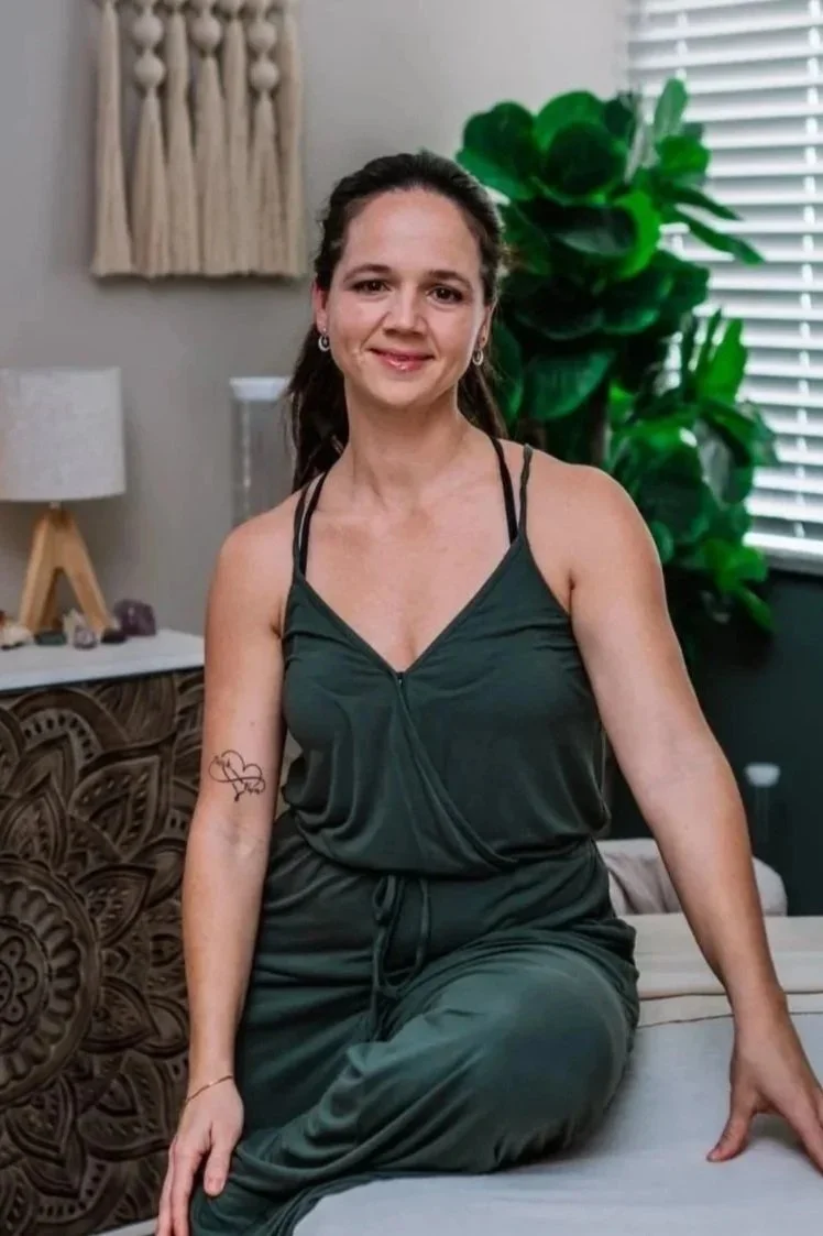 A woman with brown hair, wearing a dark green sleeveless jumpsuit, sitting on a bed in a cozy room with plants, a window with blinds, and decorative wall hangings.