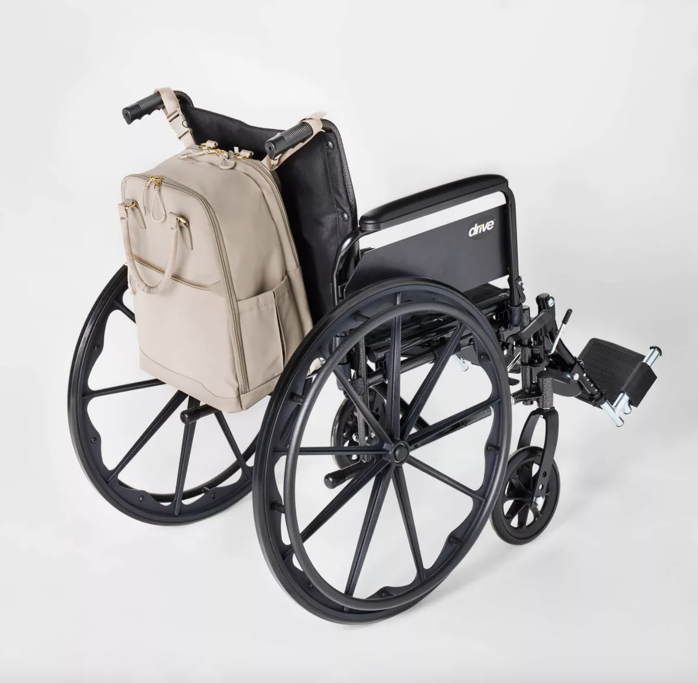 A sleek tan adaptive backpack mounted on a wheelchair via chair handle mounts.The two main compartments have heavy duty zippers with gold hardware, there is a trolley strap, and side pockets for water