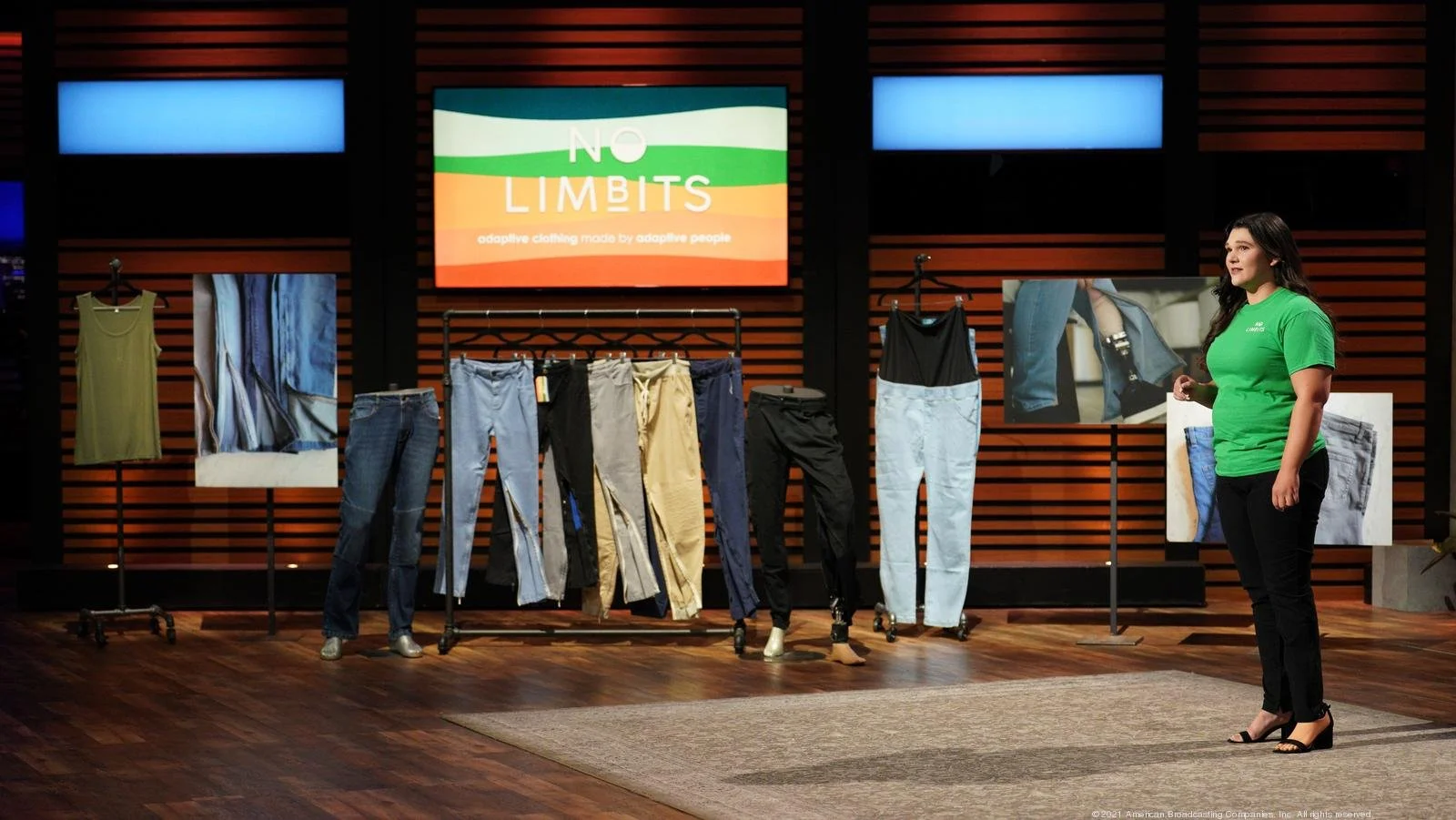 No Limbits founder Erica Cole dressed in a green t-shirt, black pants and black heels standing on the set of Shark Tank about to pitch. There is a screen with her logo, and a rack with her amputee pants in light wash, dark wash, black, and khaki