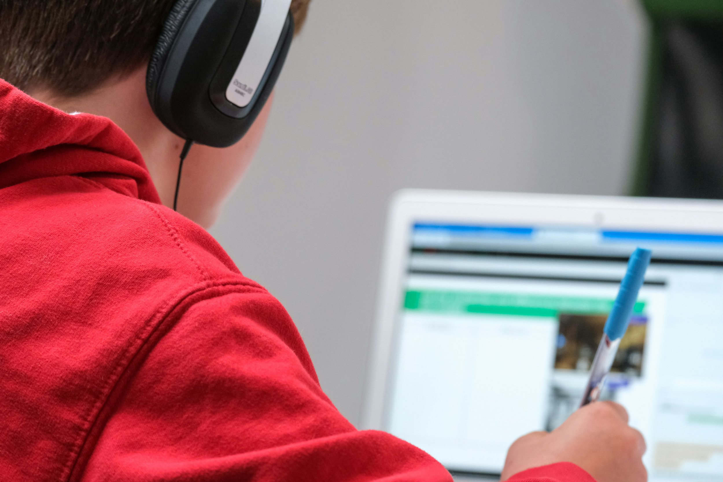 A photo from behind a young boy (around 8) with brown hair and a red sweatshirt wearing headphones. He is holding a pen and looking at something on the computer in front of him.