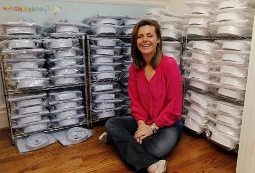 A picture of Maura Horton, founder of MagnaReady, sitting on the floor in front of stacks of adaptive, magnetic button down mens shirts smiling at the camera