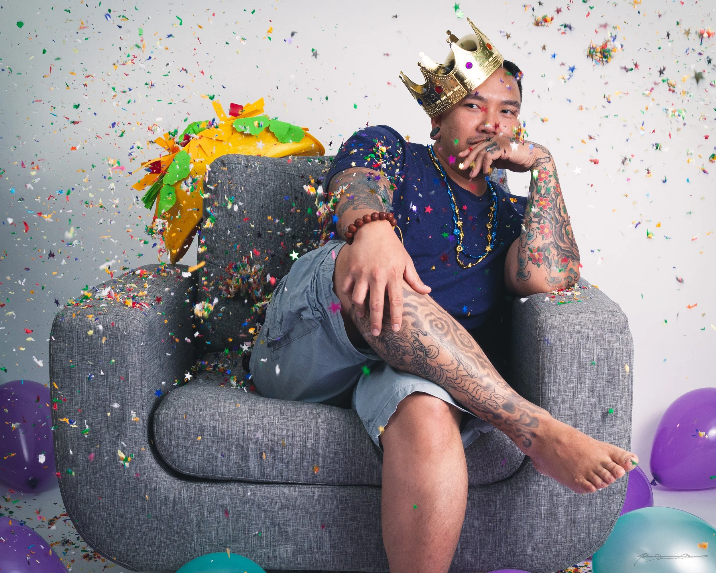 A tattooed man wearing a crown, dark blue shirt, gray shorts, sitting on a gray armchair surrounded by colorful balloons and confetti, with a yellow party hat on the chair's backrest.