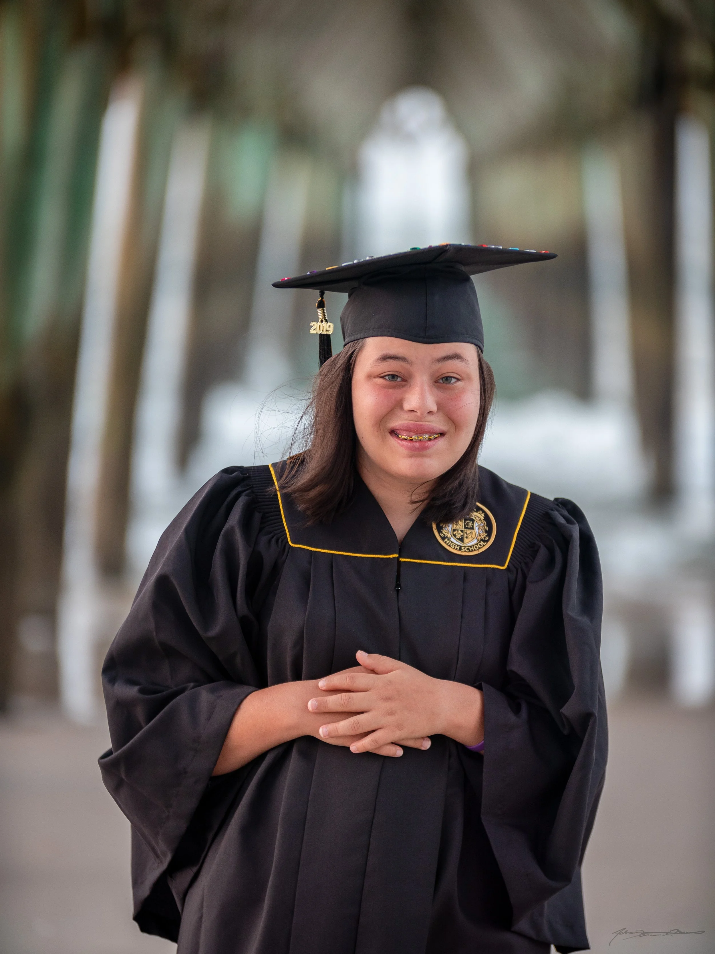 A young woman in a graduation gown and cap, with a gold tassel marked 2019, standing outdoors with trees in the background.