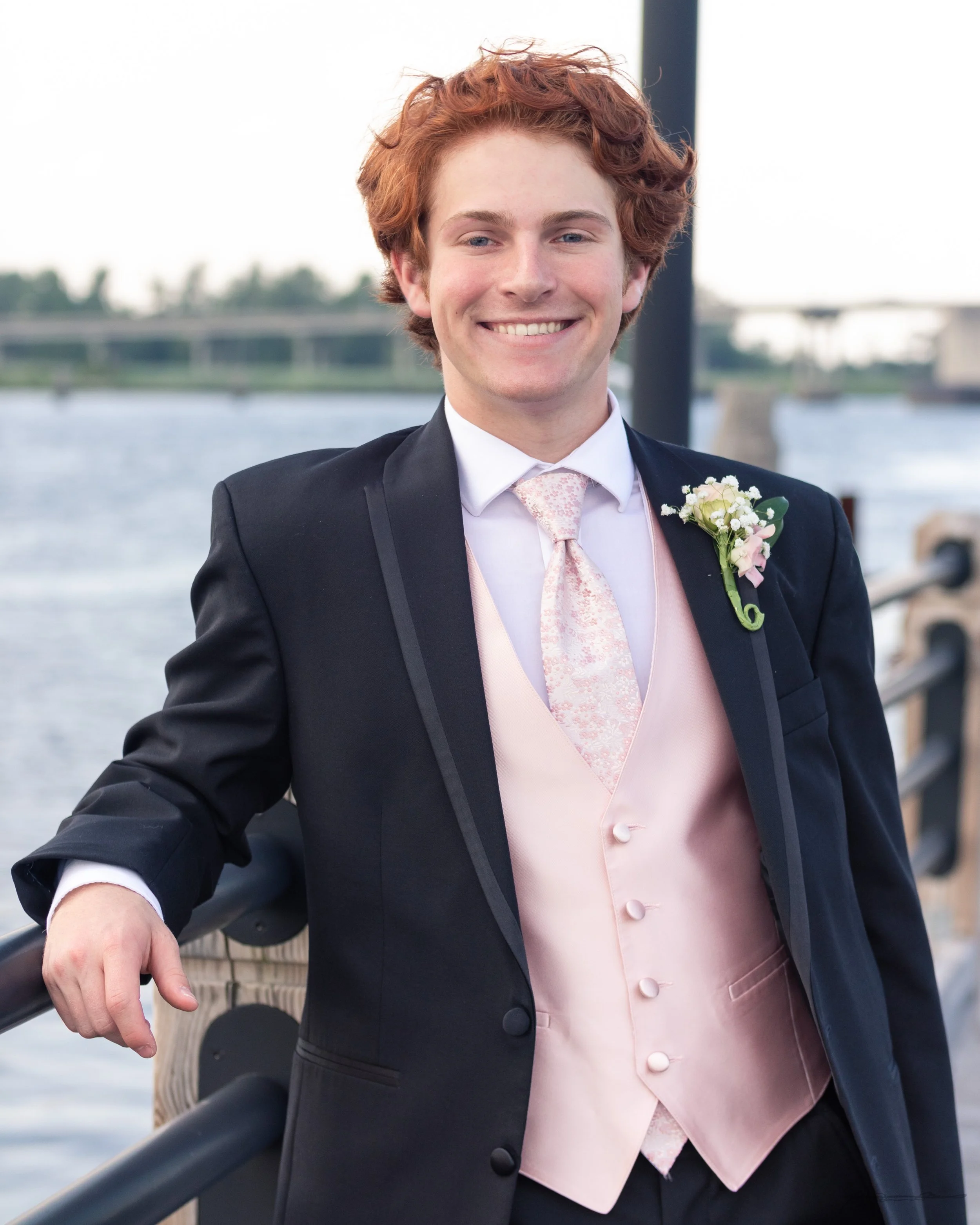 A young man in formal attire smiling outdoors near a body of water, wearing a black suit jacket, a pink waistcoat, a white shirt, and a light pink tie, with a boutonniere on his left lapel.