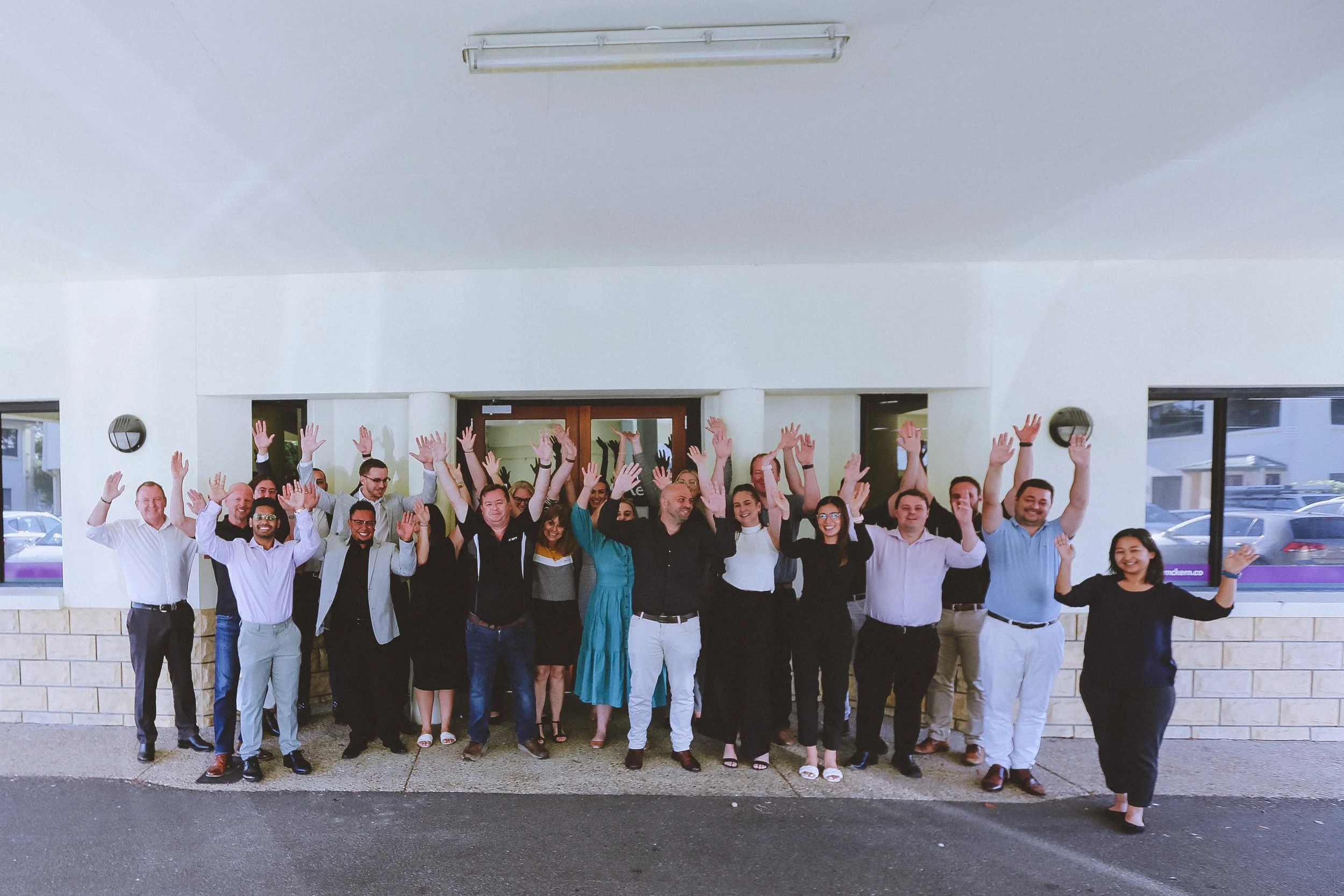 Group of diverse professionals posing inside a building with white walls and large windows, raising their hands and smiling.