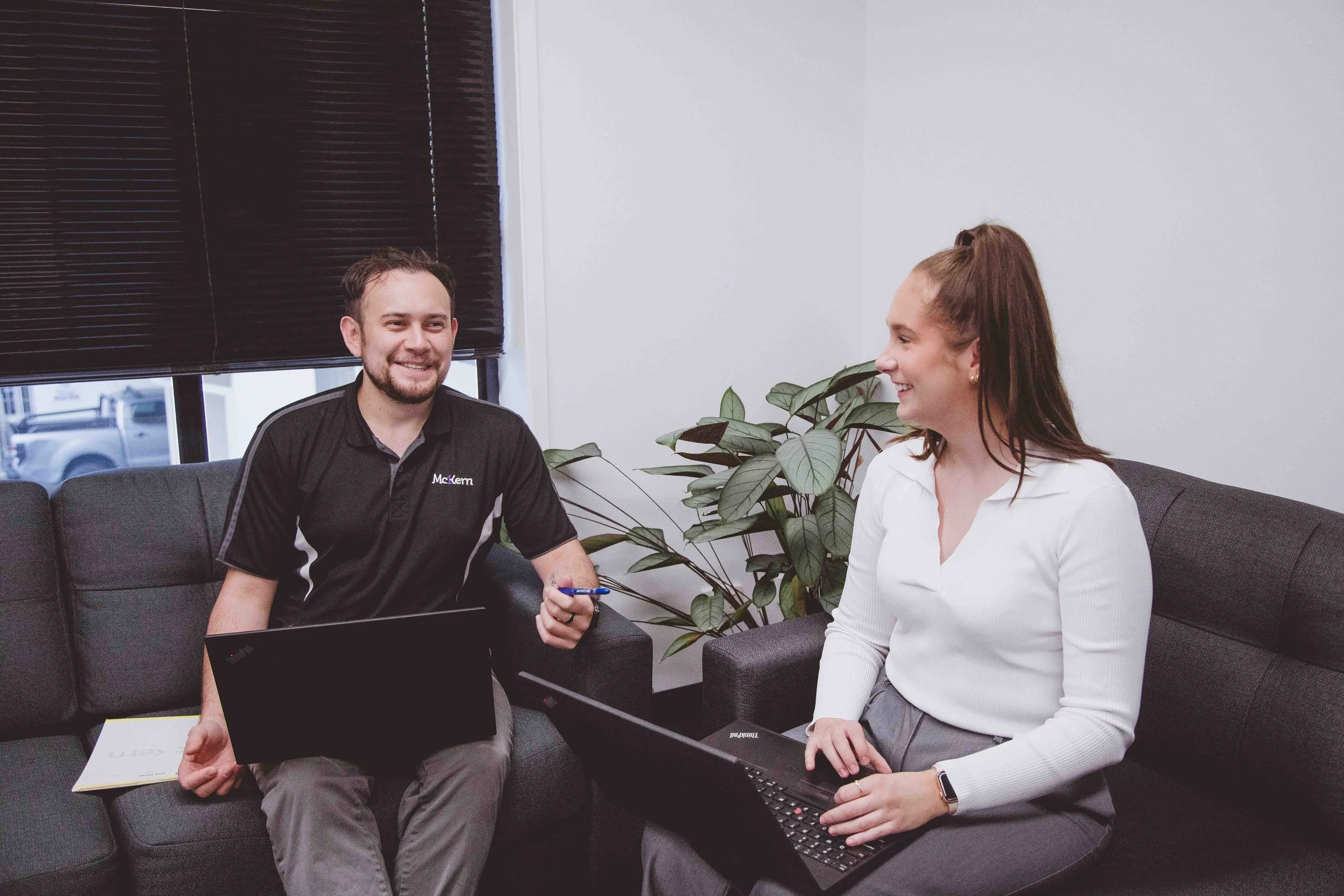 A man and woman sitting on gray couches in an office, smiling and conversing with each other. Both have laptops on their laps, and there are some papers beside the man. The background has a large plant and a window with closed blinds.