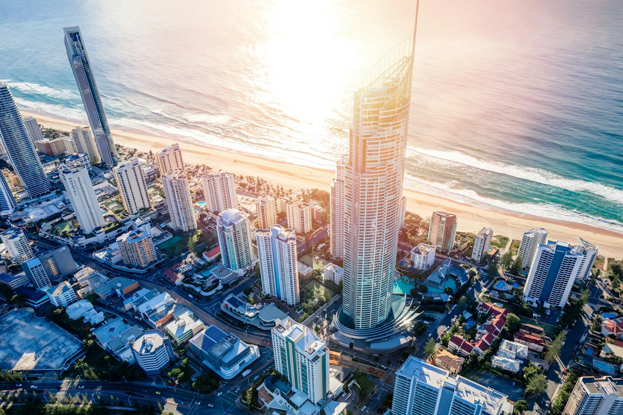 Aerial view of a coastal city with a skyline of modern skyscrapers, sandy beaches, and the ocean in the background during sunset.