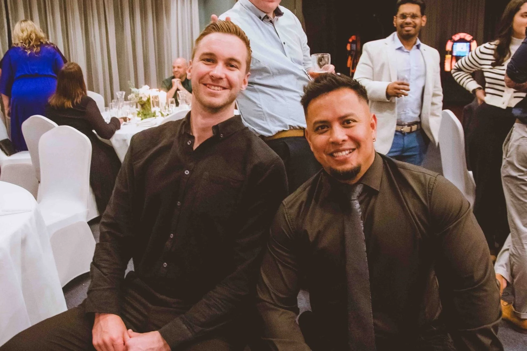 Two smiling men sitting together at a formal event, surrounded by other guests in a decorated reception hall with tables and curtains.