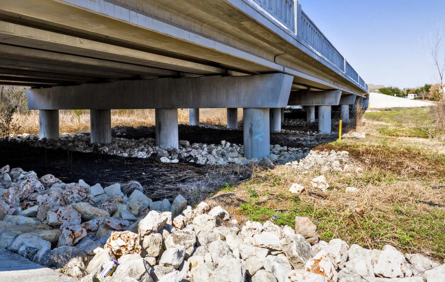 View of the underside of a bridge supported by concrete pillars, with rocks and dry grass underneath.