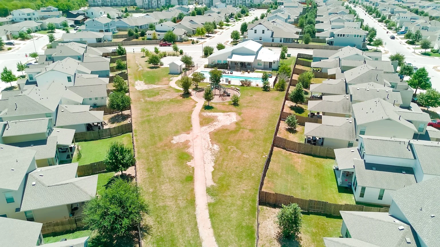 Aerial view of a residential neighborhood with a large, fenced green space featuring a walking path, a playground, and a swimming pool, surrounded by houses and streets.