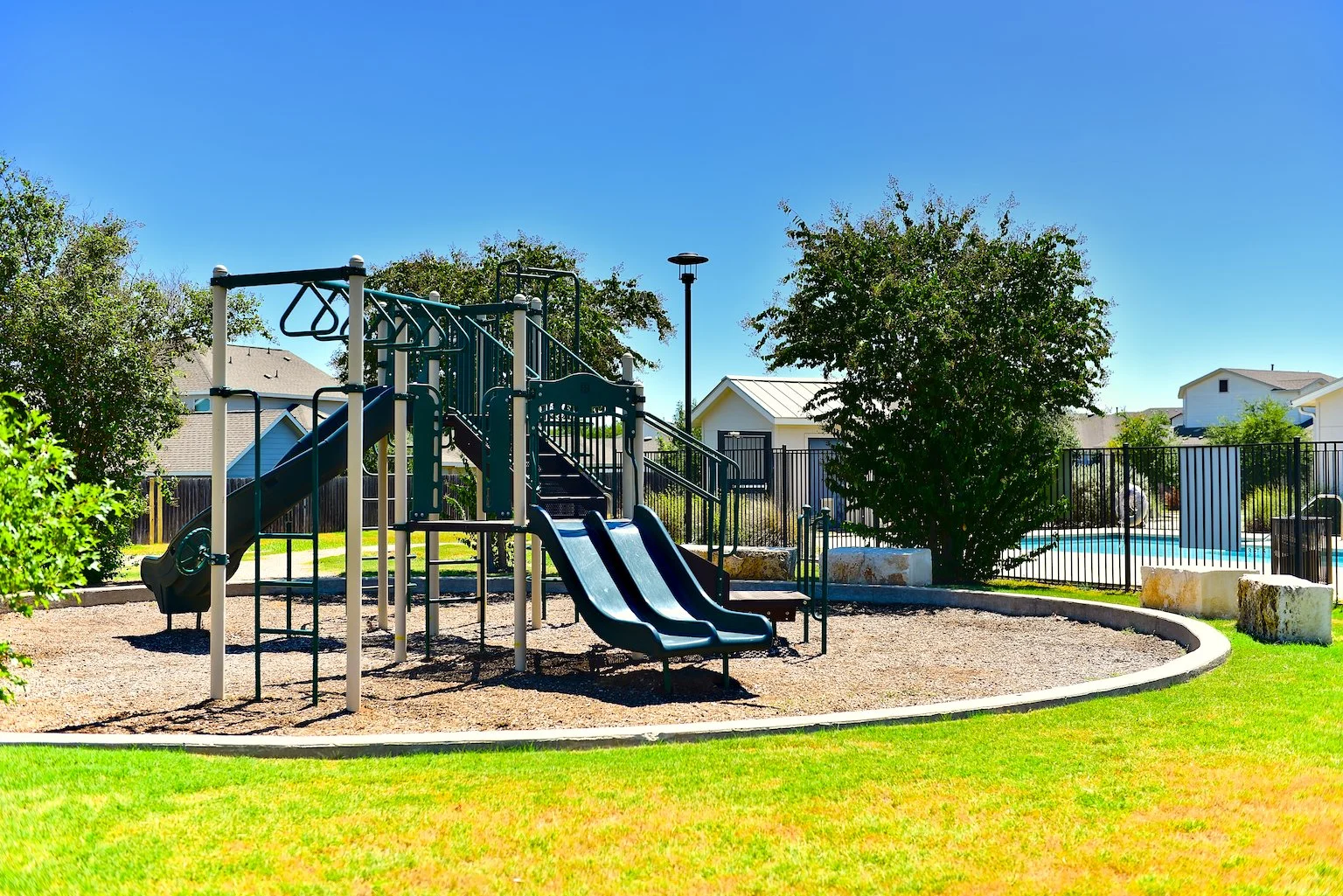 A playground with blue slides and climbing equipment in a park, surrounded by grass and trees, with residential houses and a pool in the background under a clear blue sky.