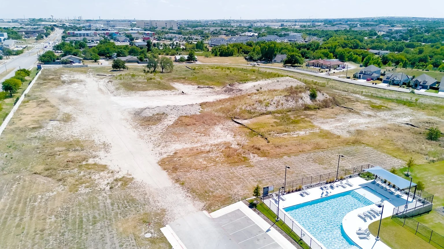 Aerial view of a vacant lot with dry grass and dirt, adjacent to a community pool with lounge chairs and fencing, in a suburban neighborhood with houses and trees in the background.