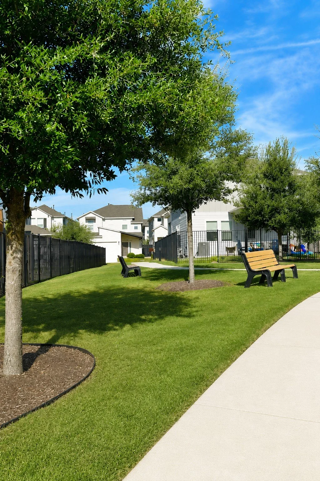 A small park or green space with benches, trees, and a grassy area. There is a sidewalk on the right side and residential buildings in the background, under a blue sky.