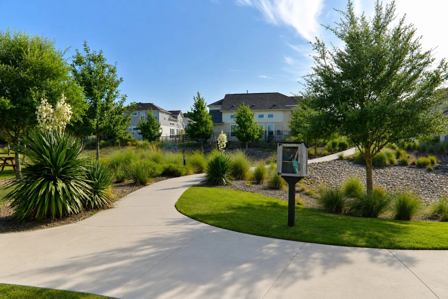 A winding concrete pathway through a landscaped yard with green grass, shrubs, and small trees. There is a glass-enclosed display case on a stand along the path. Modern houses can be seen in the background under a clear blue sky.
