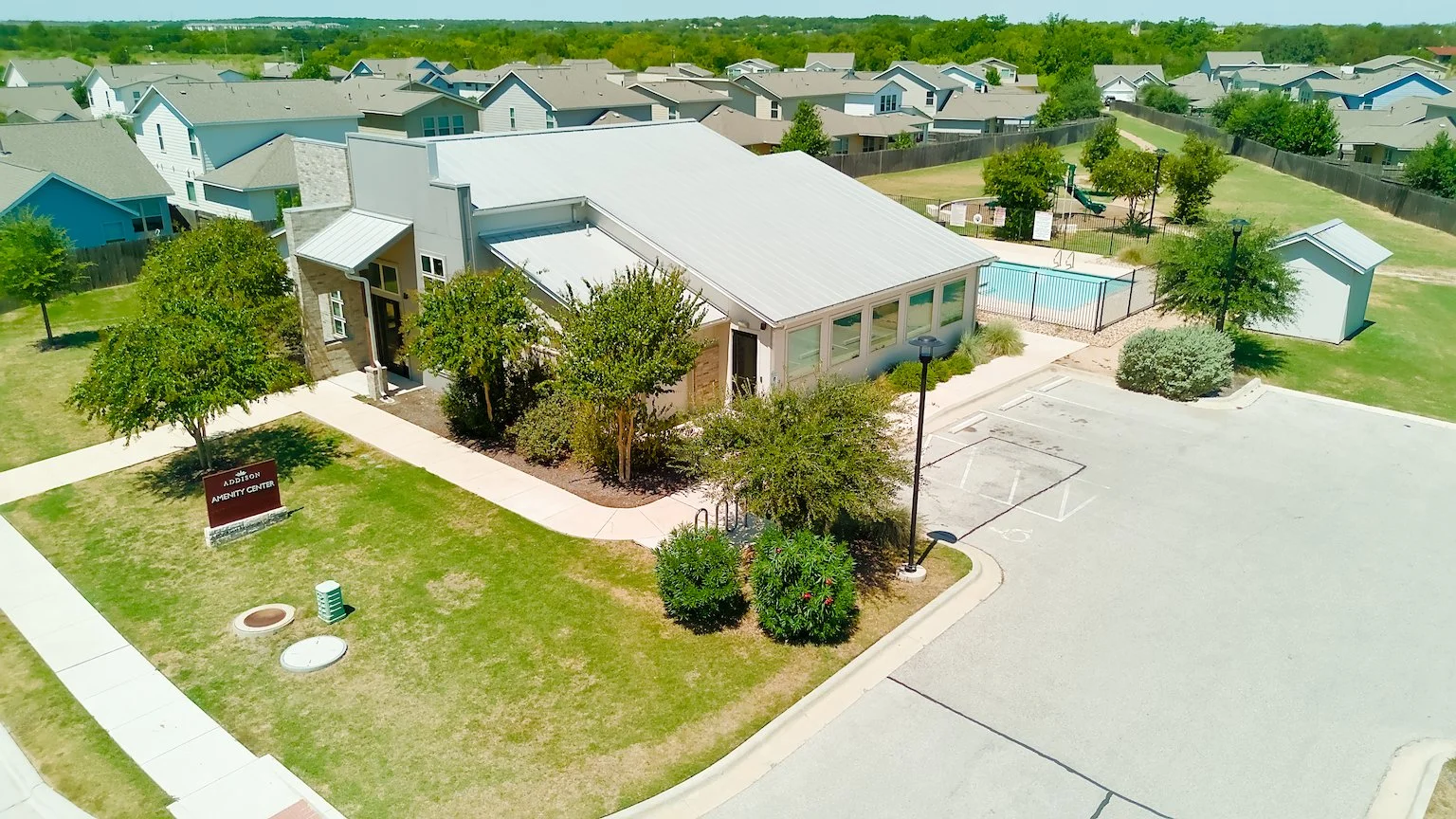 Aerial view of a community recreation area with a building, a parking lot, a swimming pool, and surrounding trees.