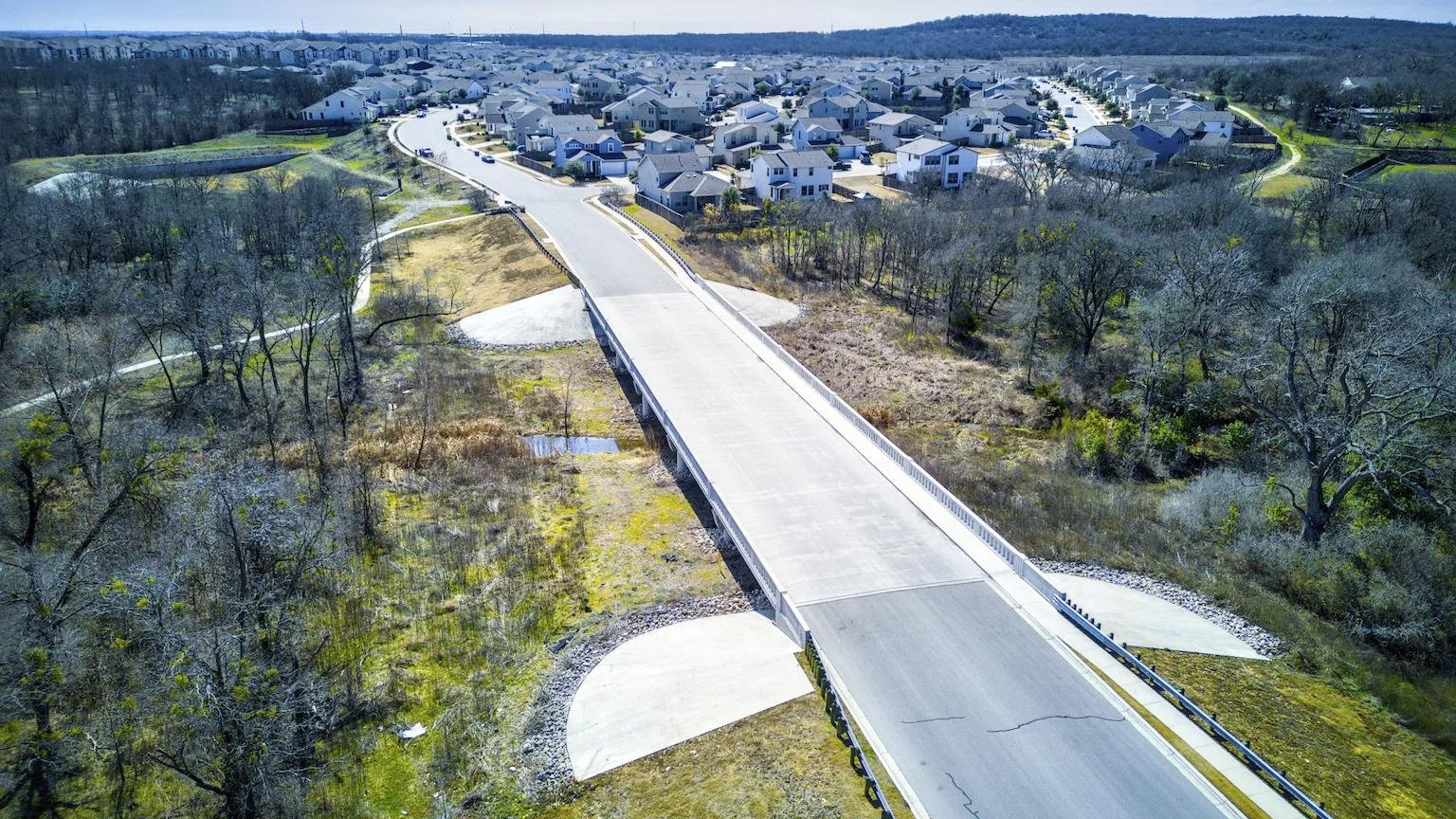 Aerial view of a new concrete bridge over a small waterway, leading to a residential neighborhood with houses and trees.