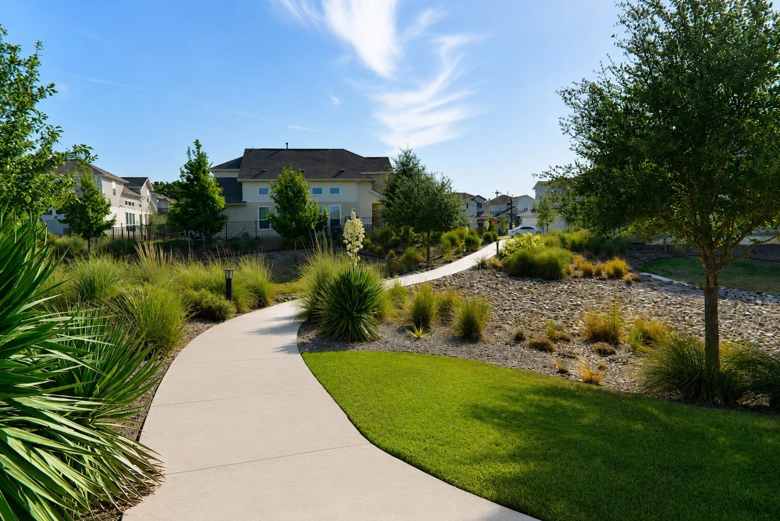 A winding concrete pathway through a landscaped garden with green grass, various plants, and trees, leading to residential houses in the background under a blue sky with a few clouds.