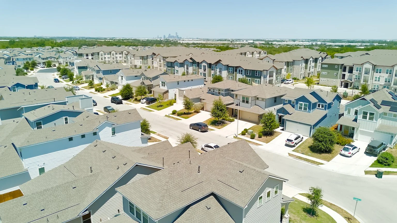 An aerial view of a suburban neighborhood with multiple modern houses, streets, cars parked outside, and some trees. In the distance, a city skyline is visible.