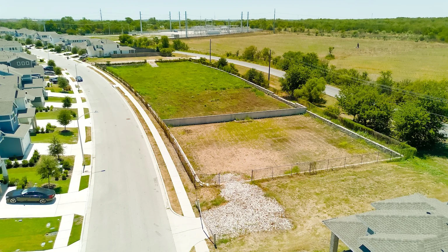 Aerial view of a residential area with new houses on the left, a road with parked cars, and vacant lots on the right, some fenced and some with patches of grass and gravel.