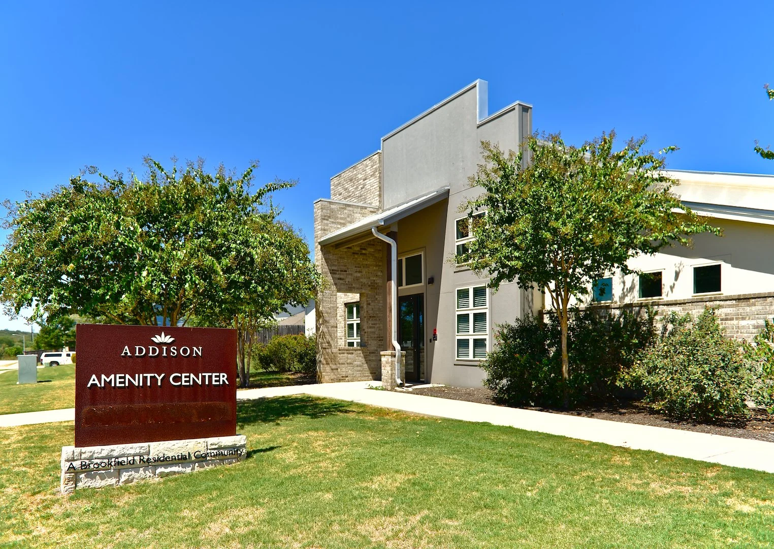A modern residential building with a sign that reads 'Addison Amenity Center' in front of two trees and a well-maintained lawn on a sunny day.